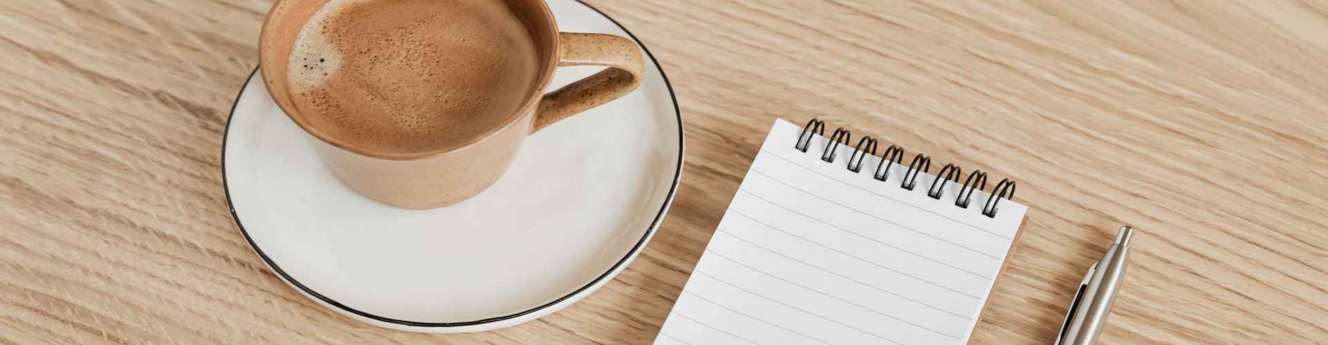 A cup of coffee, a spiral notebook and a pen on an oak desk